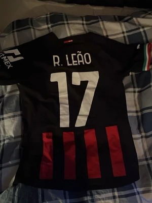 AC Milan matchtröja R. Leão #17 Puma - Snygg svart AC Milan matchtröja från Puma med R. Leão och nummer 17 på ryggen. Tröjan har röda vertikala ränder, italiensk flagga på ärmen och officiell Puma Authentic-märkning. Perfekt för fotbollsfans som vill sticka ut på läktaren eller planen.