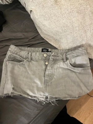 Grå jeansshorts från 157 - Snygga grå jeansshorts från 157 med råa, fransiga kanter och klassisk femficksdesign. Shortsen har bälteshällor, knappgylf och en avslappnad passform. Perfekta för varma dagar och enkel att matcha med allt.