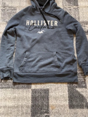 Blå hoodie från Hollister California - Snygg blå hoodie från Hollister California med vit och svart text på bröstet. Klassisk känguruficka framtill, ribbade muddar och justerbar huva med svarta snören. Mjuk insida för extra komfort, perfekt för chill dagar.