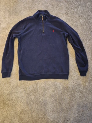 Blå halvzip tröja från Polo Ralph Lauren - Mörkblå långärmad tröja från Polo Ralph Lauren med halvzip och röd broderad logga på bröstet. Tröjan har ribbade muddar och krage, perfekt för en avslappnad men stilren look. Tröjan har däremot små tecken på användning vid högra bröstet men det har aldrig varit ett problem.