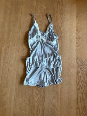 Silvrig body med spetsdetaljer - Säljer en silvrig body med tunna axelband och v-ringning. Bodyn har vita spetsdetaljer över bysten och är gjord i ett glansigt, mjukt material som känns lyxigt mot huden. Perfekt för dig som gillar en feminin och elegant stil.