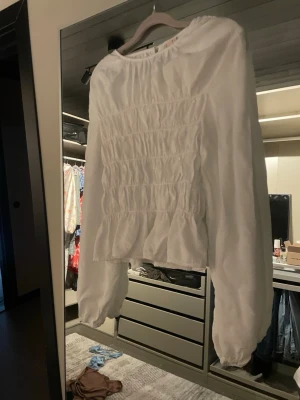 Vit blus med smock och volang - Trendig vit blus med smockdetaljer över bröstet och volang nedtill. Blusen har rund halsringning och långa puffiga ärmar med resår vid handlederna. Perfekt att styla till jeans eller kjol för en fräsch look. Aldrig använd.