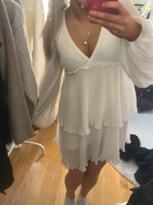 Vit omlottklänning med volanger - Säljer en söt vit kortklänning med omlottmodell och volangdetaljer. Klänningen har långärmade, lätt transparenta ärmar och v-ringad hals. Materialet känns lätt och luftigt, perfekt för varma dagar. Passformen är normal och kjolen har flera lager för extra volym.