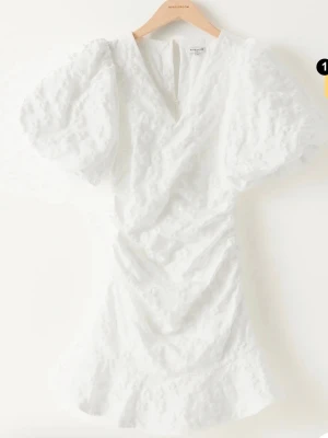 Vit omlottklänning med volang - Säljer en vit kortklänning från Bubbelroom med omlott-design, puffiga kortärmar och volang nertill. Klänningen har en v-ringad hals och är tillverkad i ett strukturerat bomullsmaterial med broderade detaljer. Perfekt för exempelvis student!