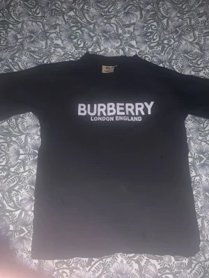 Svart Burberry t-shirt med logga - Snygg svart t-shirt från Burberry med stor vit logga 'BURBERRY LONDON ENGLAND' på bröstet. Klassisk passform och rund halsringning, perfekt för dig som gillar stilrena märkesplagg.