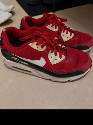 Nike Air Max röda sneakers - Snygga Nike Air Max sneakers i rött med vita och svarta detaljer samt beige partier vid snörningen. Klassisk Air Max-sula och vit swoosh på sidan. Ovandelen är i mesh och syntetmaterial för en sportig look. Perfekta för dig som gillar streetstyle.