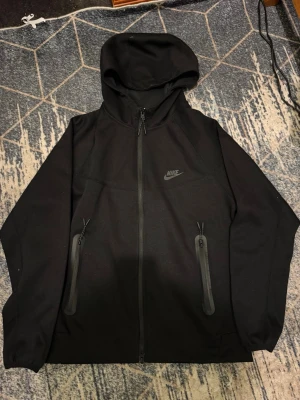 Svart Nike hoodie med dragkedja - Snygg svart hoodie från Nike med hel dragkedja framtill och två stora fickor med dragkedja. Klassisk Nike-logga på bröstet och huva för extra stil och komfort. Tillverkad i mjukt material som passar perfekt till streetwear.
