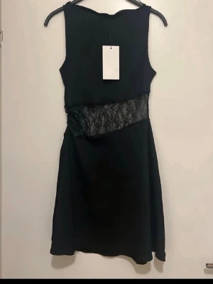 Svart klänning med spets från Zara - Snygg svart klänning från Zara med ärmlös design och transparent spetsdetalj i midjan. Kort modell som ger en modern och trendig look. Perfekt för dig som vill sticka ut med en elegant men edgy stil.