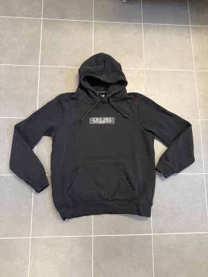 Svart hoodie från Cross Jeans stl M - Säljer en svart hoodie från Cross Jeans med logga på bröstet. Hoodien har en klassisk känguruficka framtill och justerbar huva med dragsko. Perfekt för en avslappnad och stilren look. Materialet känns mjukt och bekvämt.