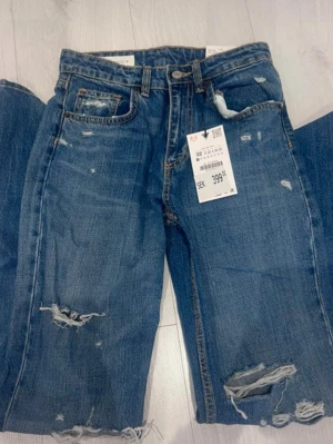 Blå slitna jeans med raka ben - Säljer ett par blå jeans med raka ben och slitningar på både lår och knän. Jeansen har klassisk femficksmodell, hög midja och råa kanter vid benslut. Perfekta för en avslappnad och trendig look. Materialet är denim i bomull.