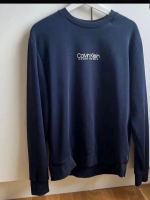 Mörkblå sweatshirt från Calvin Klein - Snygg mörkblå sweatshirt från Calvin Klein med vit logotyp på bröstet. Tröjan har rund halsringning, långa ärmar och ribbade muddar vid ärmslut och nederkant. Perfekt för en avslappnad och stilren look. Storlek L