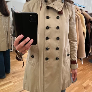 Burberry kappa med skinndetaljer  - Burberry gör ju mycket fina kappor och Trenchcoats. Den här köpte jag för rätt många år sedan i Milano. Den är använd och fodret är slitet, men lagat. Den är också kemtvättad några gånger så den är hel och ren. Jag kan träffas eller skicka den till dig om du vill. Mvh Carina Andersson 