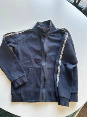 Mörkblå zip hoodie Tommy Hilfiger stl 164 - Snygg mörkblå hoodie från Tommy Hilfiger med dragkedja framtill och Hilfiger-logga på bröstet. Dekorativa band med Hilfiger-tryck längs ärmarna och två fickor fram. Tillverkad i mjuk bomullsblandning och har ribbade muddar vid ärmslut och nederkant.