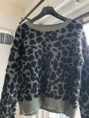 Grön leopardmönstrad stickad tröja - Stickad tröja från Vila i grönt med svart leopardmönster. Tröjan har rund halsringning, långa ärmar och ribbstickade muddar vid ärmslut och nederkant. Perfekt för dig som gillar djurmönster och vill ha en trendig look.