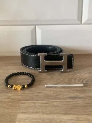 Svart skärp med stor H-spänne - Snyggt svart skärp med ett stort, silverfärgat H-spänne. Skärpet är gjort i skinn med vit kontrastsöm längs kanterna. Perfekt accessoar för att lyfta vilken outfit som helst.