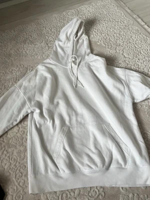 Vit oversized hoodie med huva - Snygg vit oversized hoodie med huva och känguruficka framtill. Perfekt för en avslappnad stil och enkel att matcha med andra plagg. Mjuk bomullskänsla och ribbade muddar vid ärmslut och nederkant.