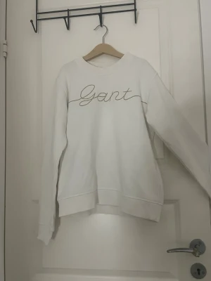 Vit sweatshirt från Gant - Säljer en stilren vit sweatshirt från Gant med broderad logga i beige framtill. Tröjan har rund halsringning, långa ärmar och ribbade muddar vid ärmslut och nederkant. Perfekt för en clean och avslappnad look.
