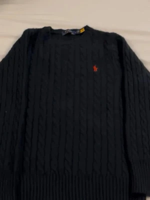 Svart kabelstickad tröja Polo Ralph Lauren - Kabelstickad svart tröja från Polo Ralph Lauren med klassisk rund halsringning och röd broderad logga på bröstet. Tröjan har långa ärmar och ribbade muddar vid ärmslut och nederkant. Perfekt för en stilren och tidlös look.