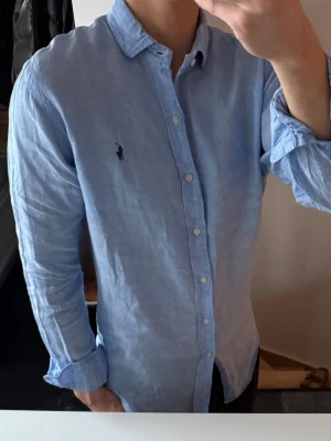 Ljusblå linneskjorta från Ralph Lauren - Säljer en stilren ljusblå skjorta från Ralph Lauren i 100% linne. Skjortan har klassisk krage, knappar framtill och den ikoniska loggan broderad på bröstet. Perfekt för en avslappnad men ändå snygg look.