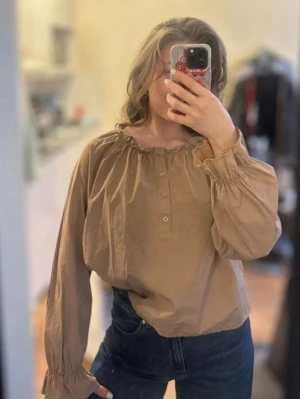 Beige blus med knappar och volang - Säljer en beige blus med rundad hals och volangdetalj. Blusen har knappar framtill och långa, puffiga ärmar med resår vid ärmslut. Materialet ser ut att vara bomull eller liknande och modellen är luftig och avslappnad.