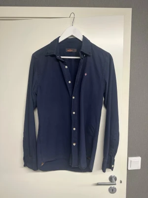 Mörkblå skjorta från Morris - Klassisk mörkblå skjorta från Morris med vita knappar och diskret logga broderad på bröstet. Skjortan har lång ärm, normal passform och är tillverkad i mjuk bomull. Perfekt till jeans eller chinos för en stilren look.