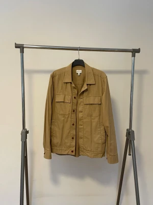 Gant Rugger Beige jeansjacka med bruna knappar - Snygg Gant rugger beige jeansjacka med klassisk krage, två bröstfickor med lock och bruna knappar framtill. Jackan har raka ärmar och en avslappnad passform som passar perfekt till många olika stilar. Pris kan diskuteras vid snabb affär.