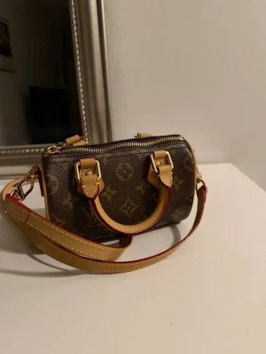 Louis Vuitton brun monogram handväska - Säljer en liten brun handväska från Louis Vuitton med klassiskt monogrammönster. Väskan har två handtag, avtagbar axelrem i beige läder med röda detaljer och guldfärgade metalldetaljer. Cylinderformad design med dragkedja upptill. PRIS KAN DISKUTERAS VID SNABBA KÖP