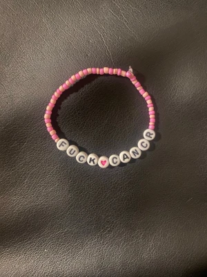 Pärlarmband med text Fuck Cancer - Handgjort armband med rosa och vita pärlor. Armbandet har bokstavspärlor som stavar ut 'FUCK CANCER' med ett rosa hjärta i mitten. Perfekt för att visa stöd och sprida ett viktigt budskap. Elastisk tråd för enkel användning.