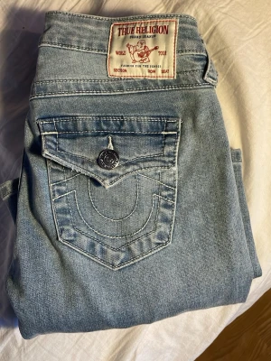 Ljusblå True Religion jeans  - Säljer ett par ljusblå jeans från True Religion, modell Joey med låg midja och bootcut. Jeansen har klassiska kontrastsömmar, snygga fickdetaljer med lock och silverfärgade knappar. Tillverkade i denim med bomullskänsla.