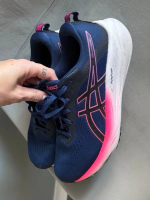 Blåa Asics sneakers med rosa detaljer - Snygga sneakers från Asics i marinblått med rosa och svarta detaljer samt vit sula. Skorna har klassisk snörning, meshmaterial för bra ventilation och en tjock, dämpande FlyteFoam-sula. Perfekta för dig som gillar sportig stil och vill ha bekväma skor till vardagen.