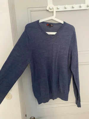 Blå merinoulltröja från Dressmann - Snygg blå stickad tröja från Dressmann i mjuk merinoull. Tröjan har V-ringning, långa ärmar och ribbade muddar vid ärmslut och nederkant. Perfekt för lager på lager och enkel att matcha med olika outfits.