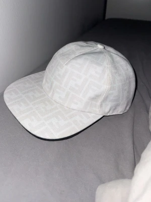Fendi keps  - Säljer en stilren vit keps från Fendi med det klassiska FF-logomönstret över hela kepsen. Kepsen har en rak skärm och är tillverkad i ett tygmaterial som känns både exklusivt och trendigt. Perfekt för dig som vill ha en snygg accessoar med lyxig känsla. 