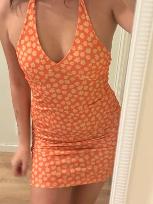 Orange blommig halterneckklänning - Snygg orange kortklänning från NA-KD x Miss Lisibell med vita blommor och halterneck. Klänningen har en djup v-ringning och figurnära passform, perfekt för sommarens alla tillfällen. Materialet känns mjukt och stretchigt.