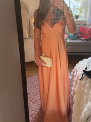Lång peachfärgad satinklänning - Säljer en lång, peachfärgad klänning i satin med smala axelband och v-ringning. Klänningen har öppen rygg med snörning och ett elegant, flytande fall. Perfekt som balklänning 