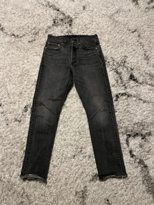Levi Jeans - Säljer ett par mörkgrå jeans med rak passform och klassisk femficksdesign. Jeansen har knappgylf och subtila slitningar för en avslappnad look. Perfekta till sneakers eller boots.