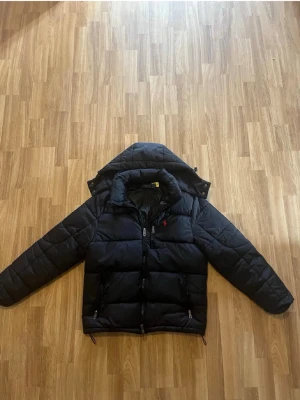 Svart pufferjacka från Polo Ralph Lauren - Snygg svart pufferjacka från Polo Ralph Lauren med avtagbar huva och tvåvägsdragkedja. Jackan har två framfickor med dragkedja och den klassiska röda Polo-loggan på bröstet. Perfekt för kalla vinterdagar och riktigt skön att bära.