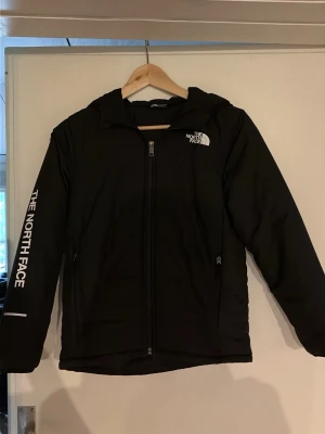Svart vindjacka från The North Face - Snygg svart vindjacka från The North Face med huva och dragkedja framtill. Logga på bröstet och ärmen, samt praktiska sidofickor. Tillverkad i ett lätt och vindtätt material som passar perfekt för blåsiga dagar.