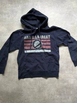 Mörkblå Saint Laurent hoodie - Snygg mörkblå hoodie från Saint Laurent med tryck framtill i vitt och rött. Klassisk huva med dragsko och magficka. Tillverkad i mjukt material som känns skönt mot huden. Perfekt för dig som vill ha en stilren och trendig look.