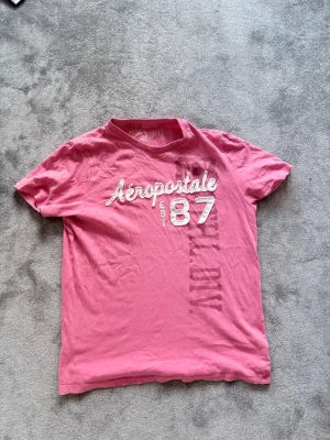Rosa Aeropostale t-shirt med tryck - Säljer en rosa t-shirt från Aeropostale med vit och ljusrosa texttryck på bröstet. Klassisk rund halsringning och korta ärmar. Perfekt för dig som gillar en avslappnad och sportig stil.
