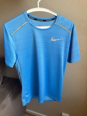 Blå Nike Dri-FIT löpartröja - Snygg blå löpartröja från Nike med Dri-FIT-material som andas och håller dig torr under träningen. Tröjan har korta ärmar, rund halsringning och reflekterande detaljer vid axlarna. Perfekt för löpning eller gympass.