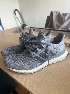 Grå Adidas Ultra Boost sneakers - Snygga grå Adidas Ultra Boost sneakers med stickad ovandel i mesh för bra andningsförmåga. Skorna har de klassiska tre ränderna på sidan, Boost-mellansula för extra komfort och svart yttersula. Perfekta för dig som gillar sportig och clean stil.