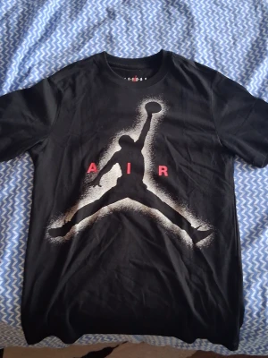 Svart t-shirt med stor Air Jordan-tryck - Snygg svart t-shirt med det ikoniska Air Jordan-motivet i vitt och svart på bröstet samt röda bokstäver. Klassisk rund hals och korta ärmar. Perfekt för dig som gillar streetwear och sportig stil.