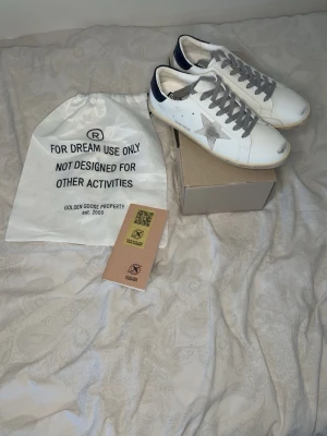 Golden Goose superstar! - Golden Goose sneakers i vitt läder med grå mockastjärna på sidan, grå skosnören och mörkblå detalj vid hälen. Klassisk låg modell med vintage-inspirerad sula och GGDB/SSTAR-tryck på sidan. Kommer med dustbag och originalkartong.