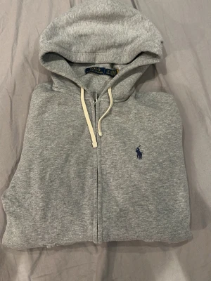 Grå zip hoodie från Polo Ralph Lauren - Säljer en grå hoodie från Polo Ralph Lauren med dragkedja. Klassisk blå logga broderad på bröstet. Mjuk bomullsblandning och avslappnad passform, perfekt för chill dagar.