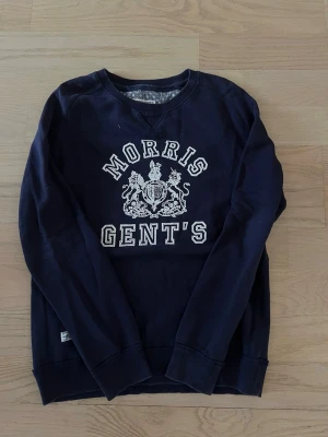 Mörkblå sweatshirt Morris GENT'S - Mörkblå sweatshirt från Morris med trycket 'MORRIS GENT'S' och ett klassiskt vapensköld-tryck på bröstet. Tröjan har rund halsringning, långa ärmar och ribbade muddar. Perfekt för en avslappnad och stilren look. Storleken är M men passar även S