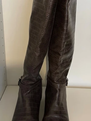 Bruna knähöga stövlar - Snygga bruna knähöga stövlar i krokopräglat skinn med låg klack och dekorativt spänne vid ankeln. Stövlarna har en stilren och trendig look som passar perfekt till höstens och vinterns outfits.