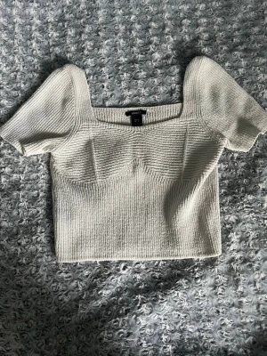 Vit stickad croppad topp - Säljer en vit stickad topp med korta ärmar och fyrkantig halsringning. Toppen är croppad och har en snygg ribbad struktur som ger en modern look. Perfekt att matcha med jeans eller kjol för en cool stil.