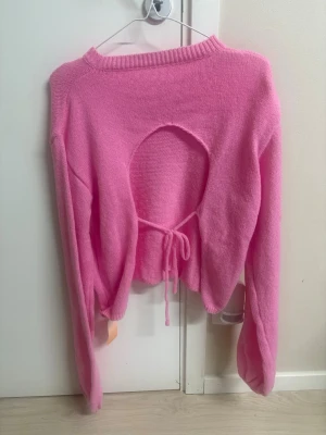 Rosa stickad tröja med cut-out - Trendig rosa stickad tröja med lång ärm och rund halsringning. Baksidan har en stor cut-out detalj och knytband i midjan för en unik look. Tröjan är croppad och har ribbade muddar vid halsen. Perfekt för dig som vill sticka ut lite extra.