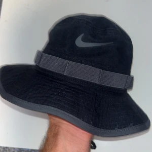 Svart bucket hat från Nike - Snygg svart bucket hat från Nike med klassisk logga framtill. Hatten har bred skärm, ventilationshål och justerbar hakrem. Tillverkad i ett tåligt tyg som ger en sportig och avslappnad vibe. Perfekt accessoar för att lyfta din streetwear-look.