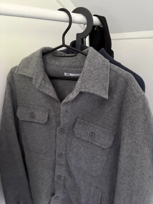 Grå overshirt från Zara - Snygg grå overshirt från Zara i mjukt, filtliknande material. Klassisk krage, två bröstfickor med lock och knappar framtill. Perfekt lager-på-lager-plagg för en avslappnad och trendig look.